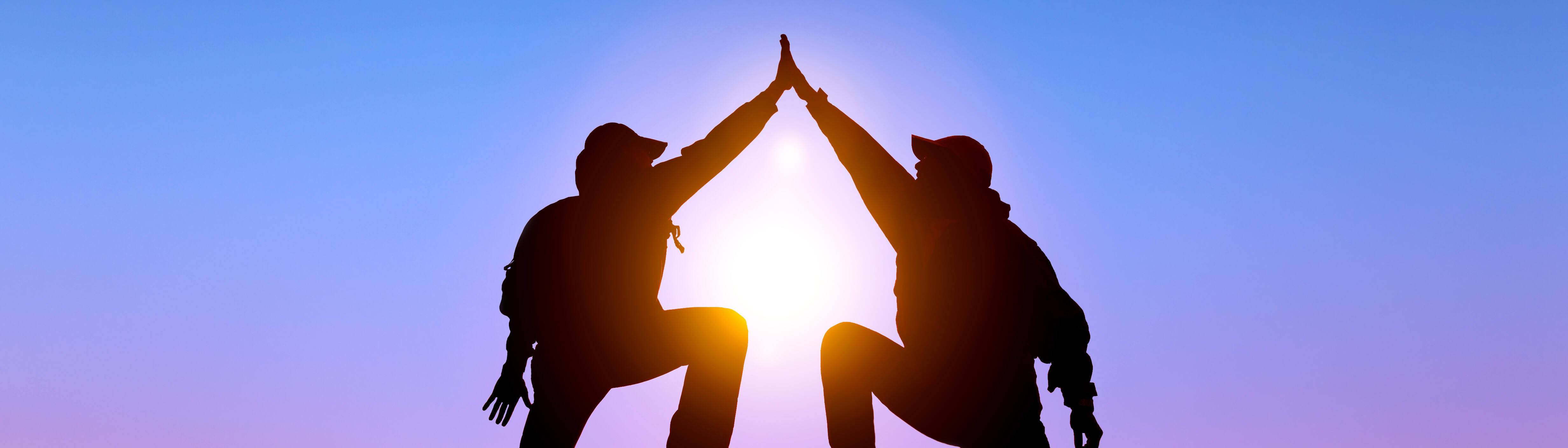 Two hikers silhouetted by the sun facing each other with arms overhead and touching the others palm